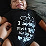 baby, smiling, onesie, black_clothing, hand, person, face, infant, lying_down, text_on_clothing, glasses_graphic, lightning_bolt, bed, blanket, skin, happy, cute, portrait, child, casual