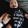 Stephen joined the competition — help win amazing prizes! baby, child, smiling, laughing, onesie, black_clothing, hand, bed, pillow, person, cute, happy, infant, face, skin, text_on_clothing, playful, tickling, indoor, closeup