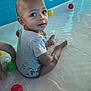 toddler, child, water, pool, plastic_balls, grey_shirt, necklace, sitting, wet_clothing, reflection, blue_wall, play, indoor_pool, curious, looking_back, barefoot, young_child, smile, water_play, portrait