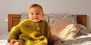 Tyam a rejoint le concours — aidez-le/la à gagner de superbes lots ! baby, bed, blanket, child, cozy, diaper, happy, headboard, indoor, mustard_sweater, patterned_bedspread, pillow, portrait, sitting, smile, socks, sunlight, toddler, wall, wooden_furniture
