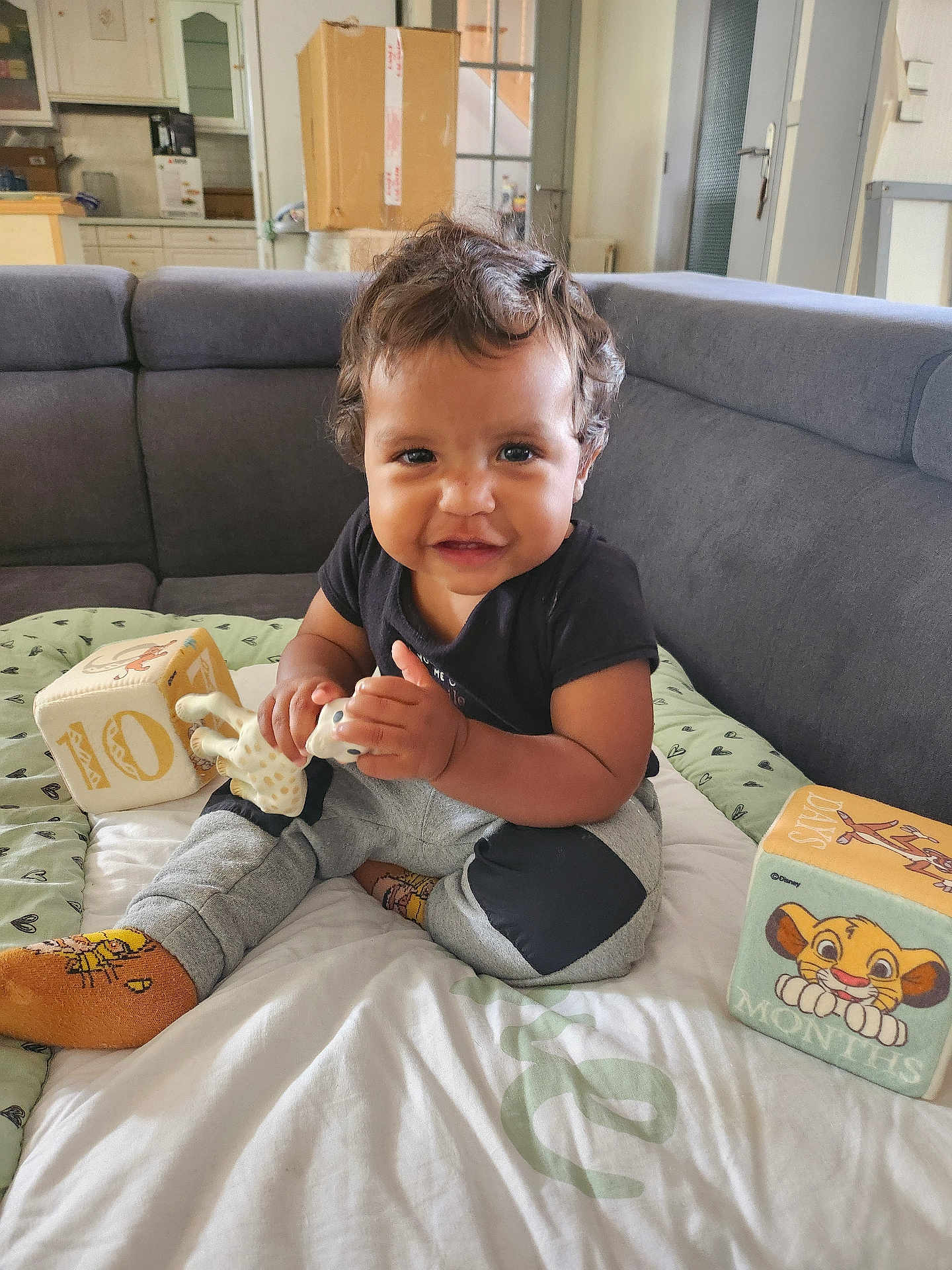Théo a rejoint le concours — aidez-le/la à gagner de superbes lots ! toddler, child, smiling, curly_hair, toy, giraffe_toy, blocks, play_mat, socks, couch, living_room, indoor, happy, sitting, clothing, person, face, home, cute, baby
