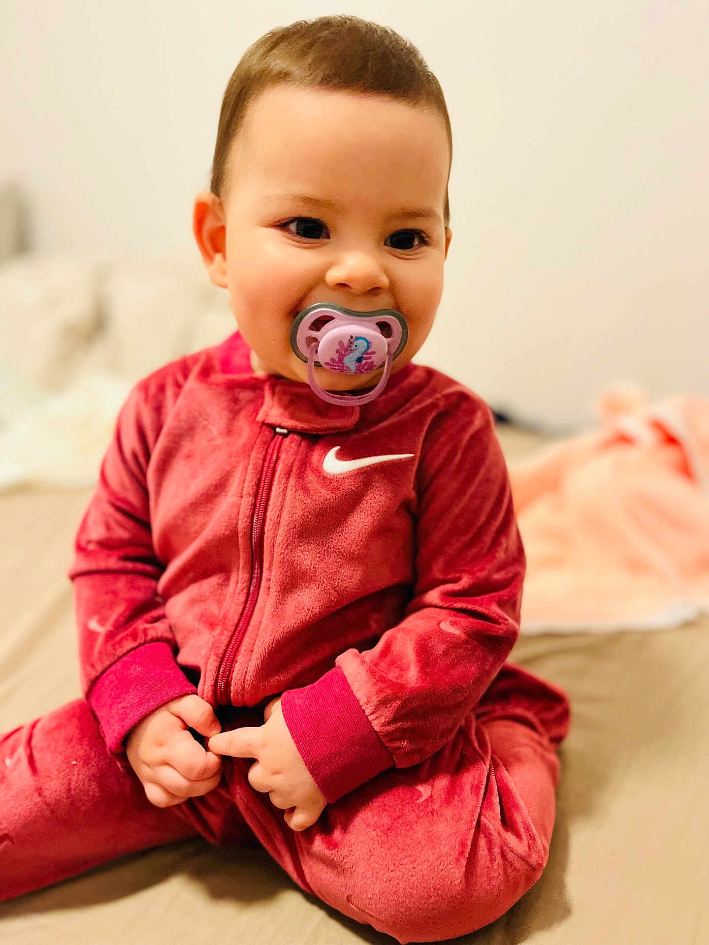 Ilinca participe au concours pour gagner de l'argent avec cette photo : baby, bed, blanket, child, cozy, cute, face, hands, happy, indoors, infant, nike_logo, onesie, pacifier, portrait, red_clothing, sitting, smiling, soft_fabric, young_child