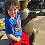 child, statue, outdoor, sunlight, smiling, blue_shirt, red_pants, concrete, bronze, happy, person, casual_clothing, daylight, standing, sculpture, playful, wristband, footwear, short_hair, expression