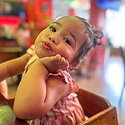 Alani is registered to the contest to win money with this photo: toddler, child, face, expression, indoor, high_chair, wood, table, person, cute, skin, hair, dress, sitting, hand, resting, blurred_background, colorful, restaurant, young