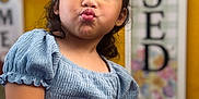 Alani is registered to the contest to win money with this photo: child, girl, blue_clothing, puff_sleeves, face, expression, duck_face, chair, wooden_chair, hand, indoor, portrait, cute, young, casual, sitting, brown_hair, skin, background_blur, yellow_background