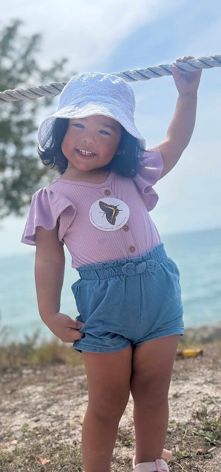 Alani is registered to the contest to win money with this photo: child, girl, smiling, hat, rope, outdoor, nature, water, shorts, sandals, grass, tree, sunny, playful, summer, portrait, happy, standing, butterfly, clothing