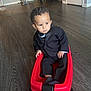 toddler, child, toy_wagon, red, black_suit, indoor, wooden_floor, serious_expression, curly_hair, person, small_wheels, flooring, household, stroller, folding_chair, door, corner, hands, socks, young_child