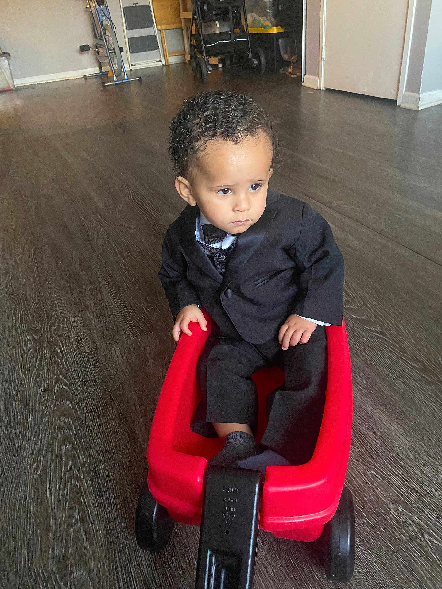 Elias joined the competition — help win amazing prizes! toddler, child, boy, suit, bow_tie, red_wagon, plastic_toy, indoor, wooden_floor, serious_expression, curly_hair, black_clothing, footwear, small_wheels, toy, person, flooring, casual_pose, home, young_child