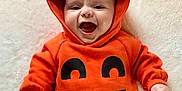 Gabriel is registered to the contest to win money with this photo: baby, infant, child, pumpkin_costume, orange_clothing, hooded_outfit, smiling, happy, cute, lying_down, face, hands, soft_surface, costume, holiday, festive, joy, expression, portrait, celebration