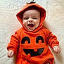 baby, infant, child, pumpkin_costume, orange_clothing, hooded_outfit, smiling, happy, cute, lying_down, face, hands, soft_surface, costume, holiday, festive, joy, expression, portrait, celebration