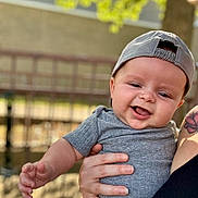 Gabriel is registered to the contest to win money with this photo: baby, child, smiling, cap, gray_shirt, tattoo, hand, outdoor, fence, sunlight, portrait, happy, person, skin, holding, casual_clothing, infant, daylight, nature, cute