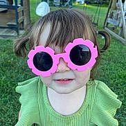 Penelope is registered to the contest to win money with this photo: toddler, child, sunglasses, pink, green_clothing, outdoor, grass, playground, play_structure, summer, cute, person, smile, hair, pigtails, face, fashion, accessory, daylight, nature