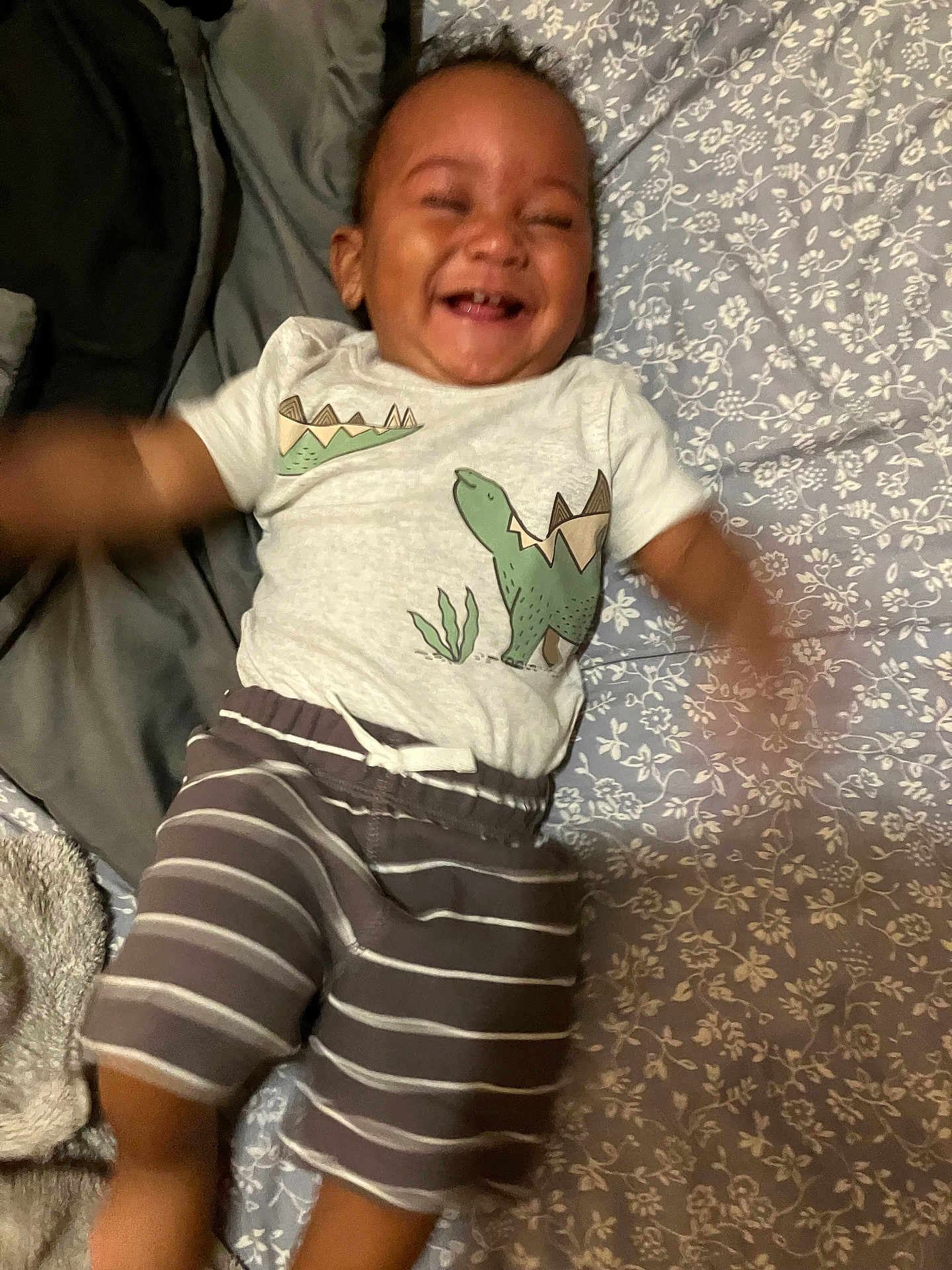 Pharaoh is registered to the contest to win money with this photo: baby, bedspread, casual, child, clothing, cute, dinosaur_shirt, floral_pattern, happy, indoor, infant, joyful, lying_down, motion_blur, person, playful, smiling, soft_fabric, striped_shorts, toothy_grin
