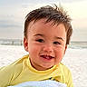 baby, child, beach, seashore, sand, ocean, water, outdoors, portrait, smiling, happy, face, head, yellow_clothing, infant, sunlight, shoreline, sea, cute, photography