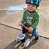 Kylan is registered to the contest to win money with this photo: toddler, helmet, dinosaur, tricycle, outdoor, sidewalk, chalk_drawing, green_sweater, blue_jeans, black_shoes, child, playground, sunny, smiling, person, toy_vehicle, safety_gear, casual_clothing, daytime, park