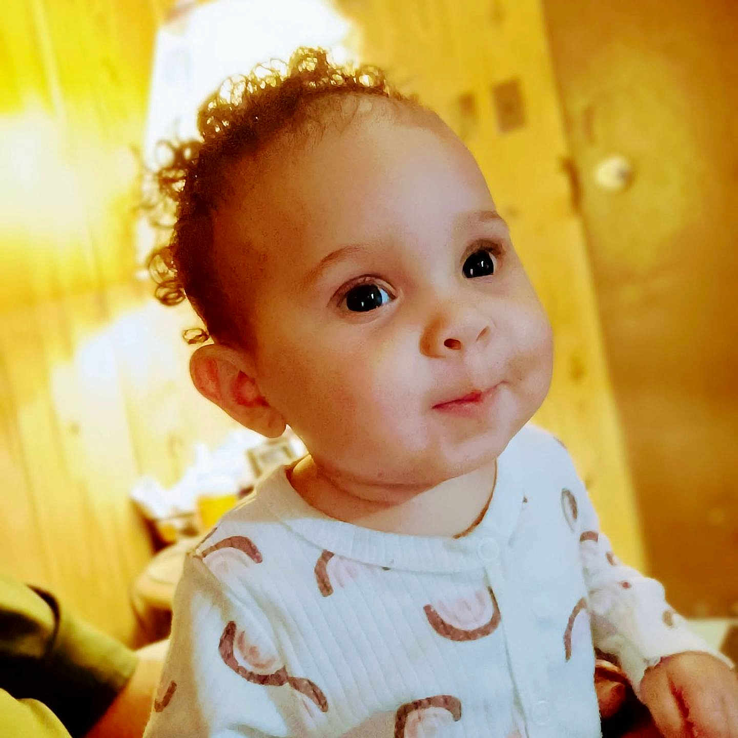 Khalani is registered to the contest to win money with this photo: baby, child, infant, portrait, face, curly_hair, smiling, happy, indoors, warm_lighting, wooden_background, home, clothing, white_outfit, patterned_clothing, sitting, close_up, photograph, adorable, human