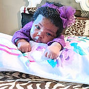 Nyomi is registered to the contest to win money with this photo: baby, infant, child, portrait, purple_outfit, hair_bow, curly_hair, lying_down, blanket, bed, bedding, zebra_print, bedroom, indoors, face, eyes, hands, looking_at_camera, colorful, adorable