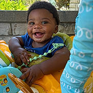 Drecoe is registered to the contest to win money with this photo: baby, smiling, child, walker, toy, outdoor, greenery, stone_wall, blue_clothing, happy, face, hands, seat, sunlight, play, infant, person, daylight, expression, cute