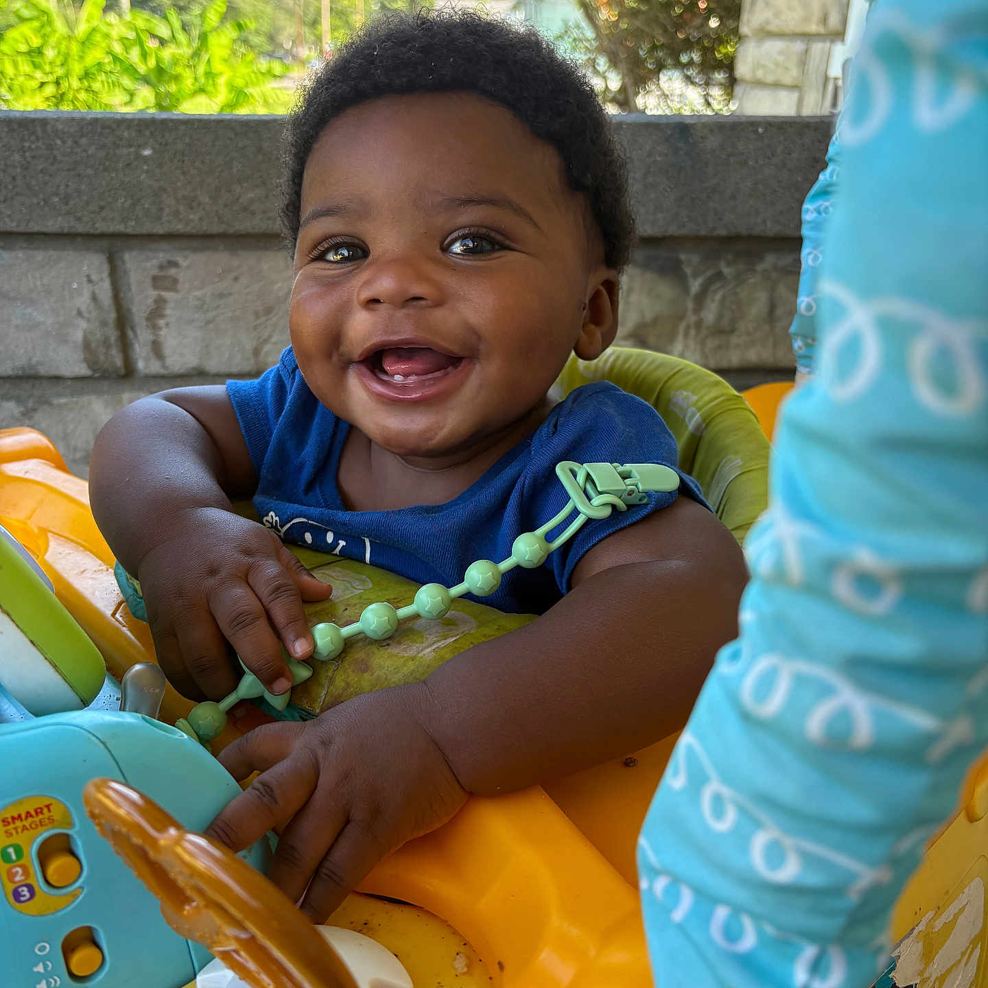 Drecoe is registered to the contest to win money with this photo: baby, smiling, child, walker, toy, outdoor, greenery, stone_wall, blue_clothing, happy, face, hands, seat, sunlight, play, infant, person, daylight, expression, cute