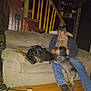 man, dog, couch, indoor, stairs, wooden_floor, beard, casual_clothing, playing, pet, sitting, furniture, home, shelf, decor, boots, jeans, canine, relaxing, interaction