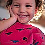 Arz is registered to the contest to win money with this photo: child, toddler, smiling, curly_hair, red_shirt, dinosaur_pattern, hand_on_face, happy, portrait, indoors, cute, young_child, person, face, bright, cheerful, casual_clothing, closeup, childhood, joy