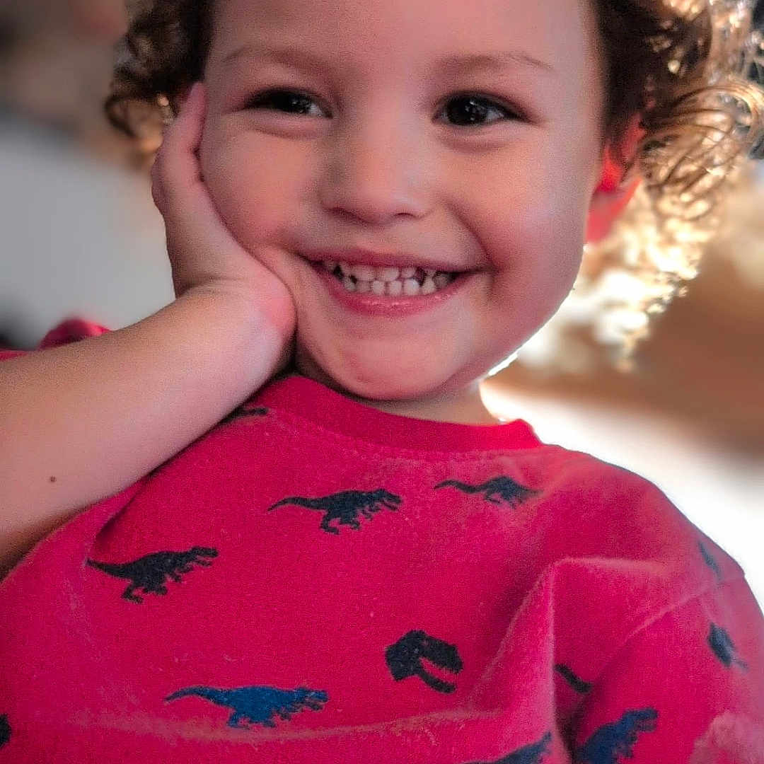 Arz is registered to the contest to win money with this photo: bright, casual_clothing, cheerful, child, childhood, closeup, curly_hair, cute, dinosaur_pattern, face, hand_on_face, happy, indoors, joy, person, portrait, red_shirt, smiling, toddler, young_child