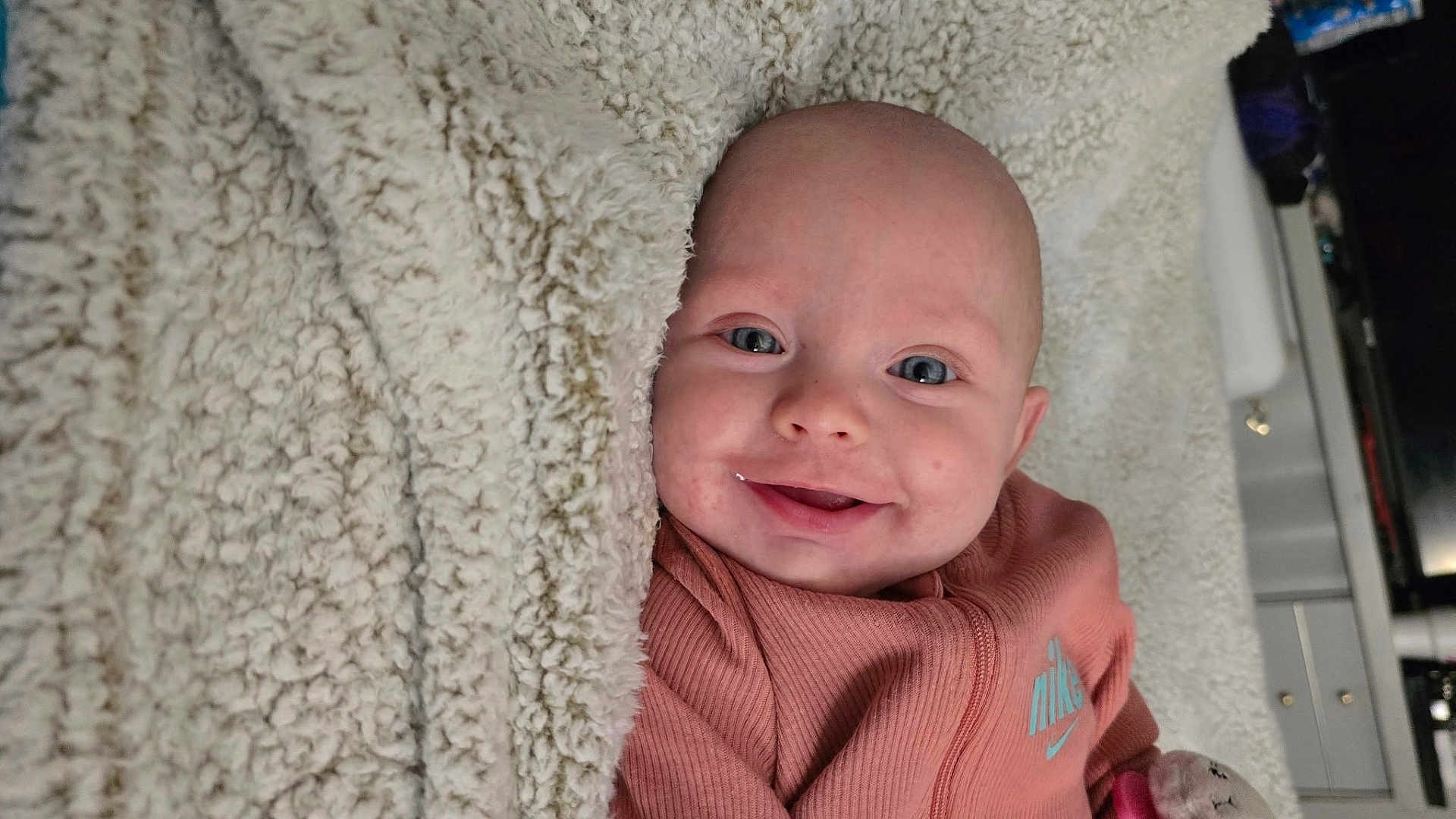 Rosalie joined the competition — help win amazing prizes! baby, smile, blanket, pink_clothing, nike_logo, drool, face, blue_eyes, infant, cozy, soft_texture, lying_down, cute, indoor, person, happy, child, head, apparel, closeup
