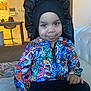 toddler, child, smile, colorful_clothing, jacket, bonnet, indoor, couch, blanket, living_room, kitchen_background, person, seated, happy, face, portrait, young_child, casual, home, cute