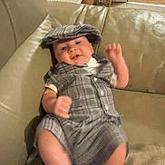 Brody is registered to the contest to win money with this photo: baby, infant, plaid_outfit, hat, couch, leather, smiling, happy, indoor, person, child, furniture, blanket, socks, living_room, cozy, home, cute, portrait, waving