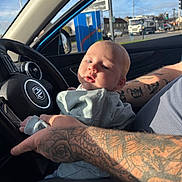 Brody is registered to the contest to win money with this photo: baby, infant, tattoo, arm, car_interior, steering_wheel, window, daylight, person, clothing, seat, hand, face, skin, vehicle, road, outside, blue_sky, casual, closeup