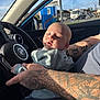 baby, infant, tattoo, arm, car_interior, steering_wheel, window, daylight, person, clothing, seat, hand, face, skin, vehicle, road, outside, blue_sky, casual, closeup