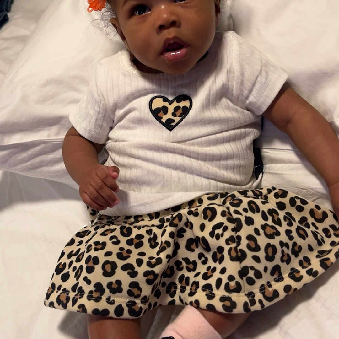 Teja joined the competition — help win amazing prizes! baby, bed, black_bows, child, clothing, curly_hair, cute, expression, hair_clips, indoor, leopard_print, orange_clips, person, pink_socks, portrait, shirt, sitting, skirt, toddler, white_bedding