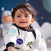 Advait is registered to the contest to win money with this photo: child, toddler, smiling, face, space_theme, astronaut_suit, indoor, cute, happy, feet, hair, blue_background, star, toy_astronaut, clothing, portrait, baby, playful, person, sitting