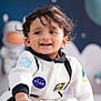 child, toddler, smiling, face, space_theme, astronaut_suit, indoor, cute, happy, feet, hair, blue_background, star, toy_astronaut, clothing, portrait, baby, playful, person, sitting