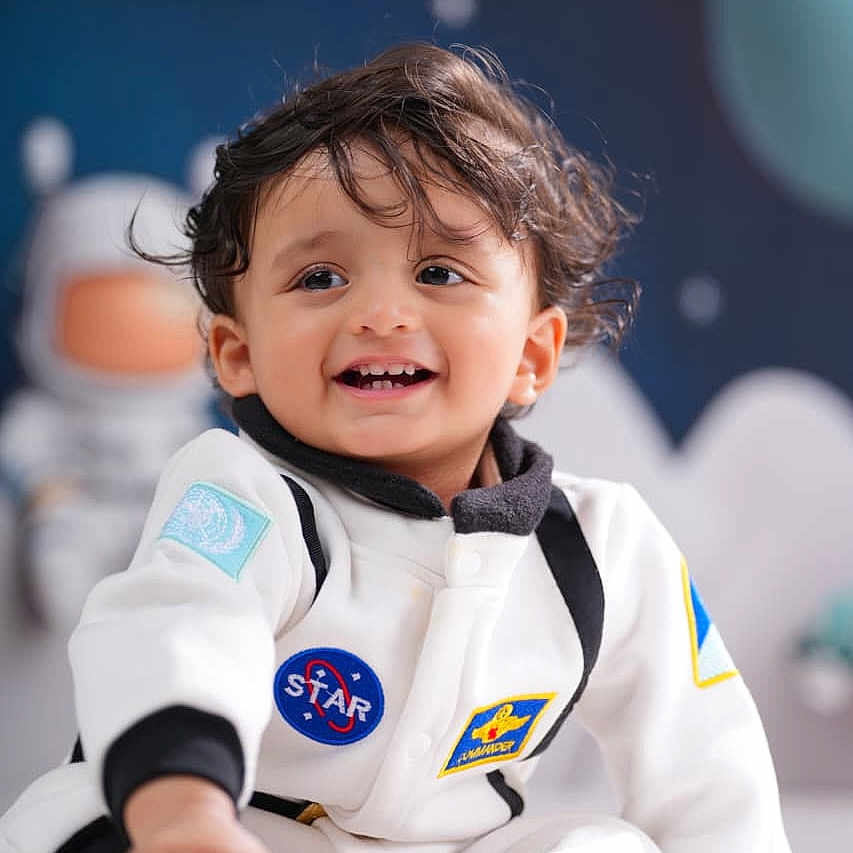 Advait is registered to the contest to win money with this photo: astronaut_suit, baby, blue_background, child, clothing, cute, face, feet, hair, happy, indoor, person, playful, portrait, sitting, smiling, space_theme, star, toddler, toy_astronaut