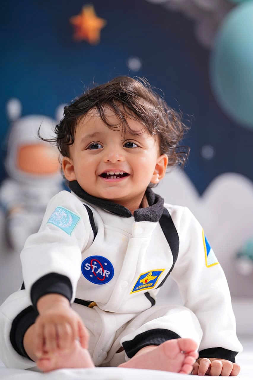 Advait is registered to the contest to win money with this photo: child, toddler, smiling, face, space_theme, astronaut_suit, indoor, cute, happy, feet, hair, blue_background, star, toy_astronaut, clothing, portrait, baby, playful, person, sitting
