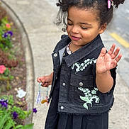 Sofya participe au concours pour gagner de l'argent avec cette photo : black_dress, casual_clothing, child, curly_hair, cute, daylight, denim_vest, embroidered_clothing, flowerbed, hand_raised, holding_objects, outdoor, pigtails, plants, portrait, sidewalk, smiling, street, toddler, young_girl