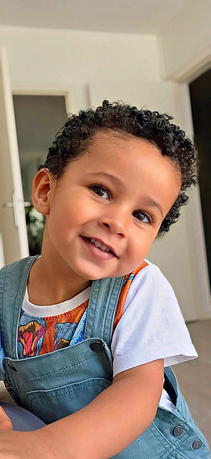 Nathaniel participe au concours pour gagner de l'argent avec cette photo : button, casual_wear, child, colorful_clothing, curly_hair, cute, dinosaur_print, eyes, face, happy, home, indoor, light_skin, overalls, person, portrait, short_sleeves, smiling, toddler, young_child
