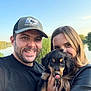 man, woman, puppy, dog, smiling, outdoor, nature, lake, trees, sky, leash, cap, black_shirt, long_hair, happy, portrait, holding, pets, couple, sunlight