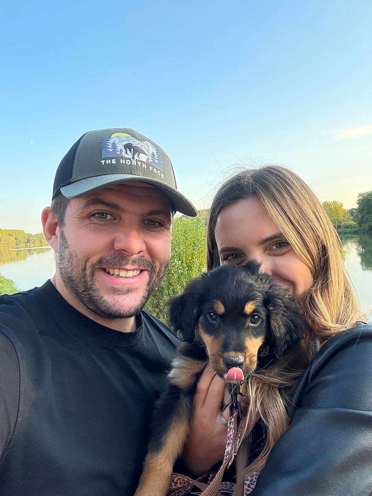 Apy participe au concours pour gagner de l'argent avec cette photo : man, woman, puppy, dog, smiling, outdoor, nature, lake, trees, sky, leash, cap, black_shirt, long_hair, happy, portrait, holding, pets, couple, sunlight