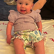 Loretta is registered to the contest to win money with this photo: baby, child, smiling, sitting, headband, bow, purple_clothing, floral_skirt, blanket, indoor, carpet, person, adult_arm, happy, cute, infant, legs, face, expression, casual