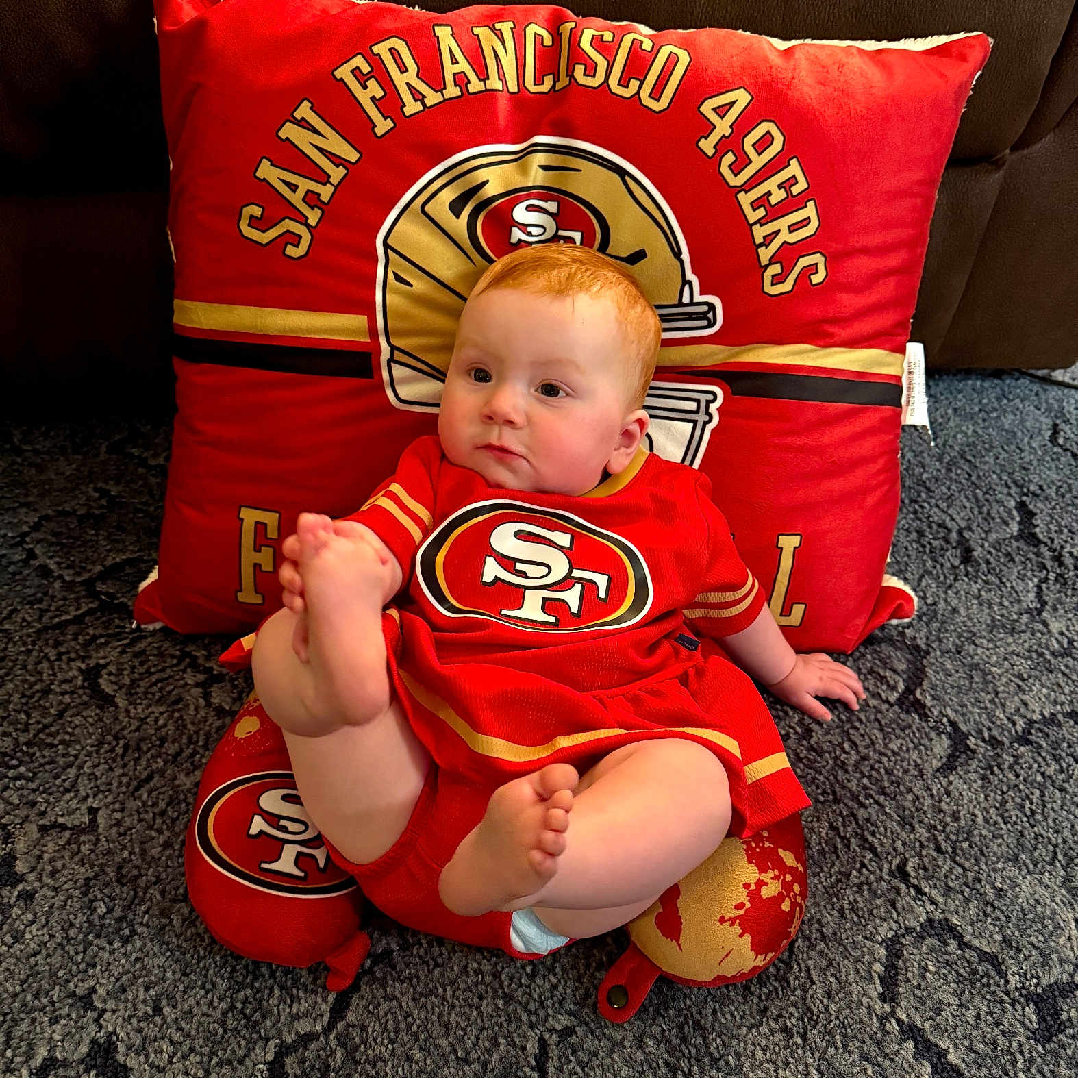 Loretta is registered to the contest to win money with this photo: baby, carpet, casual, child, couch, cute, foot, football_team_logo, home, indoor, infant, pillow, plush, portrait, red_clothing, san_francisco_49ers, sitting, soft_texture, sports_fan, team_colors