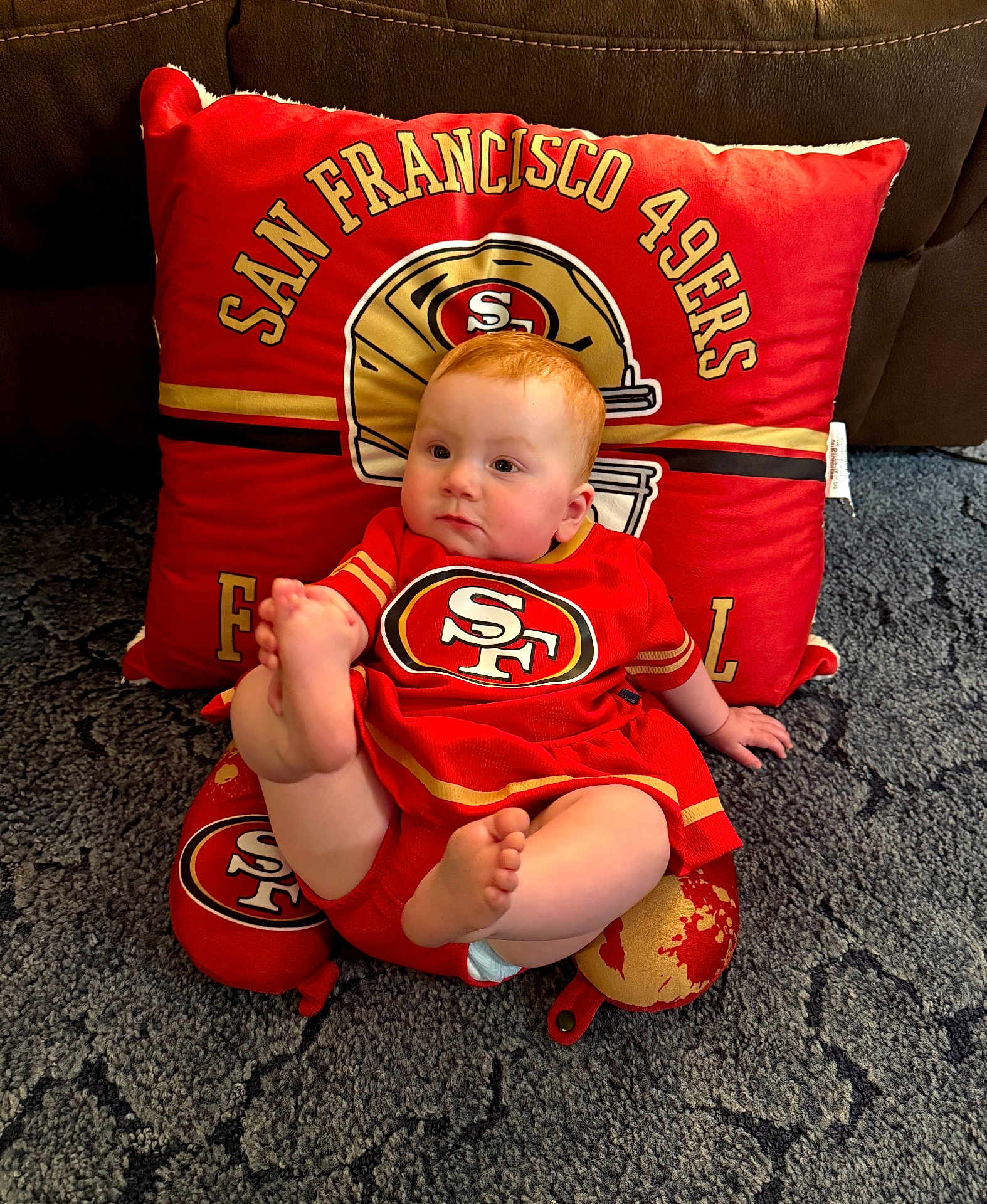Loretta is registered to the contest to win money with this photo: baby, infant, red_clothing, san_francisco_49ers, pillow, football_team_logo, carpet, indoor, cute, child, foot, sitting, sports_fan, team_colors, plush, soft_texture, couch, home, portrait, casual
