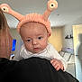 Callen is registered to the contest to win money with this photo: baby, infant, person, hat, pink_hat, antennae, face, blue_eyes, hand, white_clothing, shoulder, indoor, living_room, couch, door, shelf, soft_lighting, curious_expression, closeup, headwear