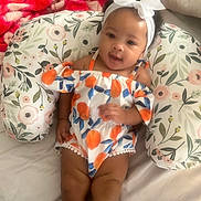 Saige is registered to the contest to win money with this photo: baby, child, infant, smiling, headband, bow, floral, pillow, blanket, bed, colorful_clothing, orange, blue, white, cute, indoors, person, portrait, happy, relaxed