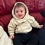 baby, infant, child, hoodie, white_hoodie, black_pants, socks, red_sofa, leather_sofa, couch, smile, wide_eyes, bright_eyes, sitting, hands, blanket, pillow, cozy, indoors, portrait