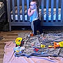 toddler, child, crib, blanket, toy, floor, indoor, wooden_floor, clothing, pants, shirt, baby_toy, stuffed_animal, play_area, curious, standing, room, home, furniture, cute
