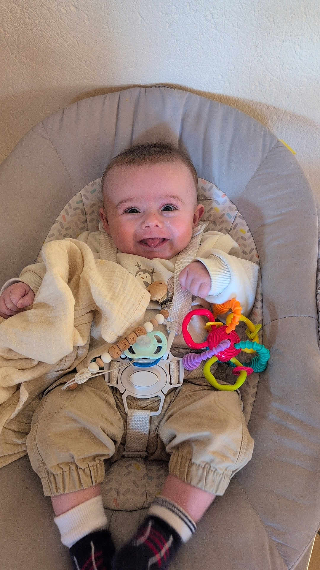 Maël a rejoint le concours — aidez-le/la à gagner de superbes lots ! baby, infant, smile, tongue_out, bouncer, toy_ring, pacifier, blanket, buckle, strap, onesie, pants, socks, cushion, chair, indoor, wall, close_up, happy, portrait