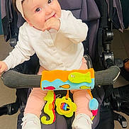 Sana participe au concours pour gagner de l'argent avec cette photo : baby, child, stroller, toy, headband, white_sweater, pink_pants, white_shoes, indoor, cute, smiling, seated, colorful_toys, playful, infant, happy, hand_in_mouth, wheel, floor, baby_accessories