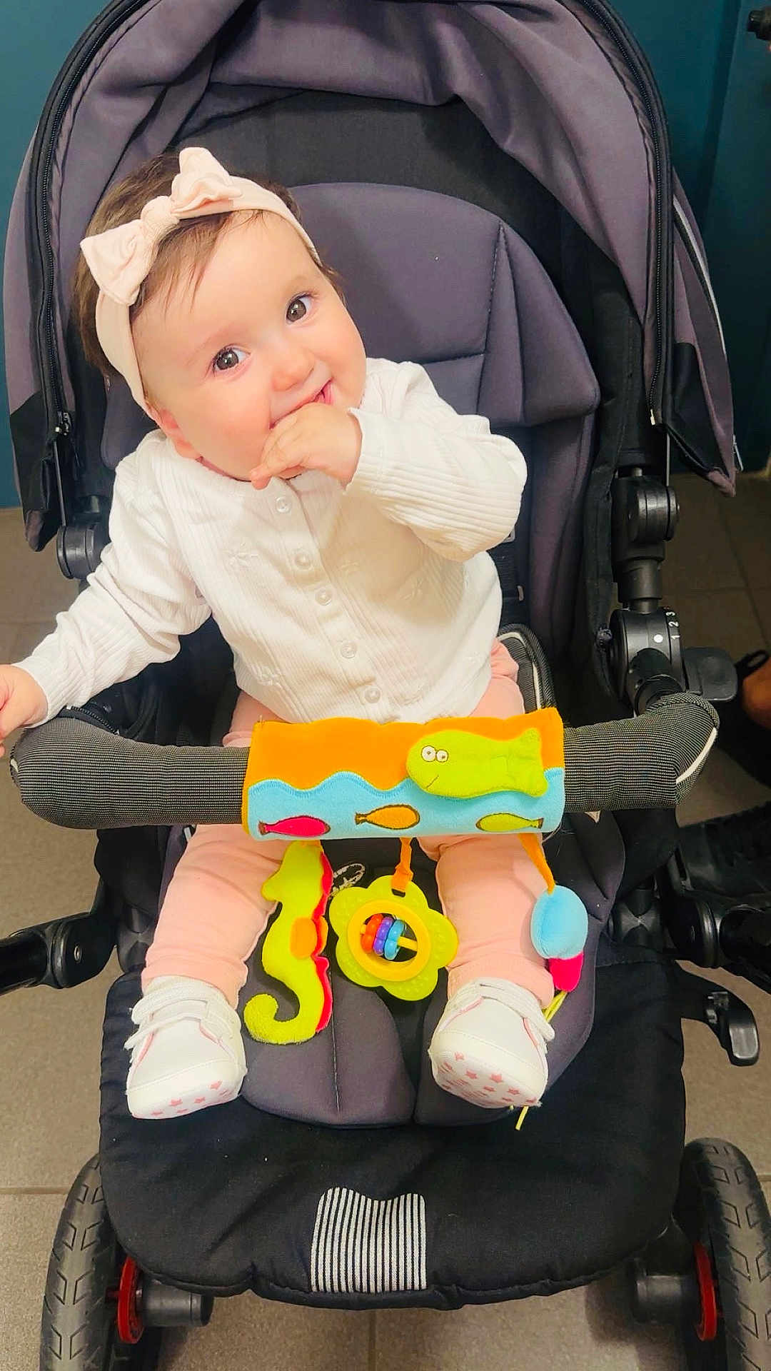Sana participe au concours pour gagner de l'argent avec cette photo : baby, child, stroller, toy, headband, white_sweater, pink_pants, white_shoes, indoor, cute, smiling, seated, colorful_toys, playful, infant, happy, hand_in_mouth, wheel, floor, baby_accessories